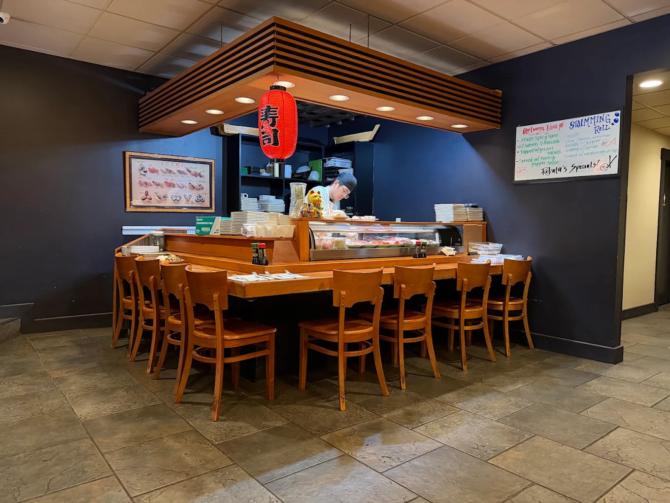 The dining room's L-shaped wooden counter with chairs, a hanging red paper lantern and a chef working behind the case