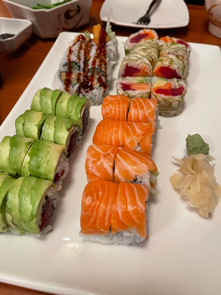 Four sushi rolls on a white platter — avocado-topped, fresh salmon, tempura with sauce and marbled seaweed rolls — with ginger and wasabi on a wooden table