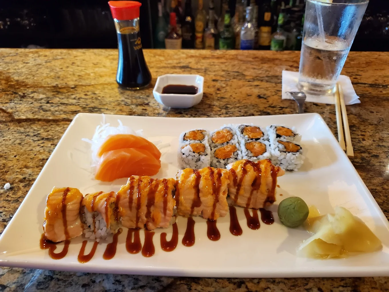 Seared salmon roll with spicy mayo and soy glaze, salmon sashimi on daikon and spicy maki on a white plate at the bar